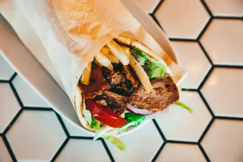 Kebab w tortilli z mięsem, frytkami, sałatą, pomidorem i cebulą na białym talerzu.
