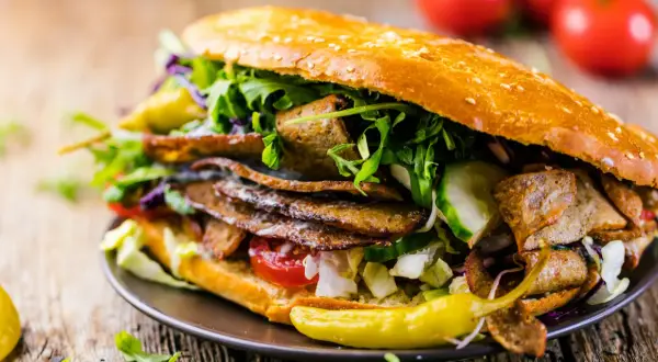 Duży kebab w bułce z mięsem, warzywami i sosem na drewnianym tle.