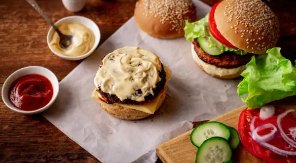 Dwa burgery, sosy (majonez, ketchup) i składniki na drewnianym stole.