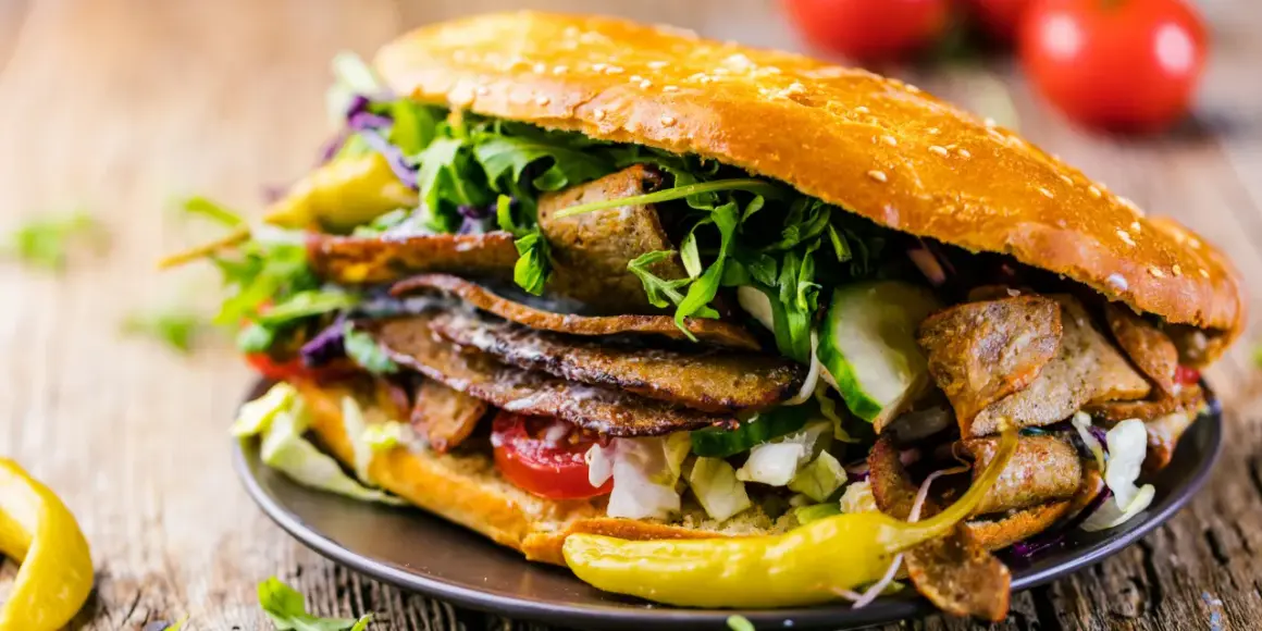 Duży kebab w bułce z mięsem, warzywami i sosem na drewnianym tle.